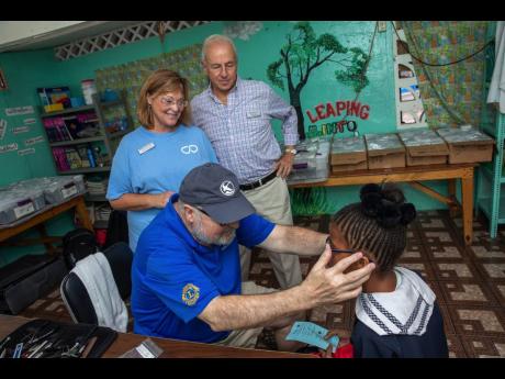 Diane Pollard, president and CEO of the Issa Trust Foundation, and Chairman Paul Issa smile as a volunteer fits a pair of glasses on a Savanna-La-Mar Primary School student.