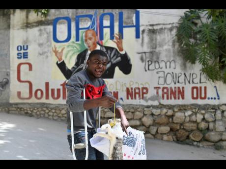 A pedestrian pulls a dead cat from a bag to scare children who were teasing him as he walks past a mural of slain President Jovenel Moise in Port-au-Prince, Haiti, on Tuesday, July 13. Moïse was assassinated on July 7 in his residence, located a few metre