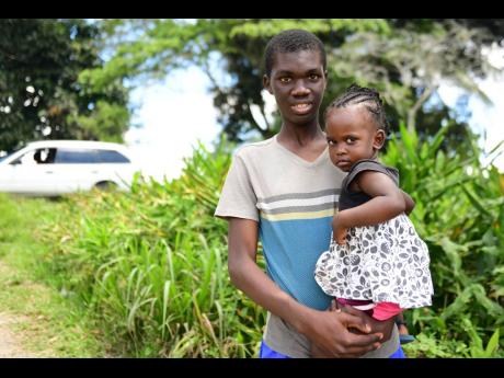 Delano Tucker is seen holding his neighbour’s child Tashaun on Mud Lane in Guy’s Hill, St Catherine. The 17-year-old has won a scholarship to attend the College of Agriculture, Science and Education.