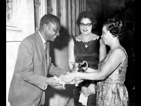 This 1964 photograph shows Allan Abrahams, retiring permanent secretary in the ministry of communications and works, is presented with a silver salver and goblet by his secretary, Sybil Wood. Mrs. Abrahams is at centre.