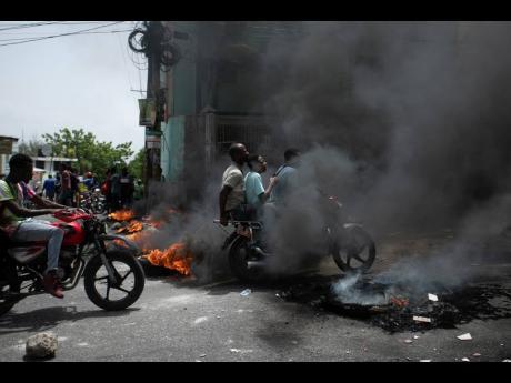 A motorcycle taxi driver carries clients past a burning barricade set by taxi drivers to protest the country’s fuel shortage in Port-au-Prince, Haiti, on Wednesday, July 13.