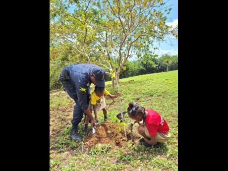 A policer joins two Golden Grove Basic School students in planting a seedling during the main National Tree Planting Day ceremony held at the Jamaica Bauxite Mining Limited Protocol House in St Ann.