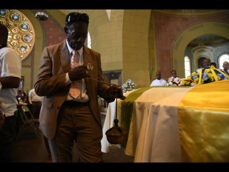 Father Kingsley Asphall (hidden behind) guides Paul Stevens, father of O’Neil Stevens, while he incenses the coffins during the funeral service for the former principal of Jessie Ripoll Primary School and his wife, personal banker Camesha Lindsay-Stevens
