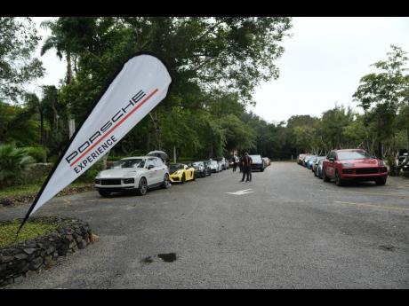 A fleet of Porsche vehicles lines a paved road surrounded by lush greenery, with a prominent Porsche Experience flag marking the event.