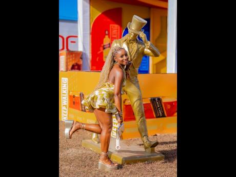 A jolt of golden glamour. Entrepreneur Ashia Hamilton lights up Rise and Toast in a tailored golden yellow jacquard set and wedge heels, striking a confident pose beside the iconic Johnnie Walker striding man.