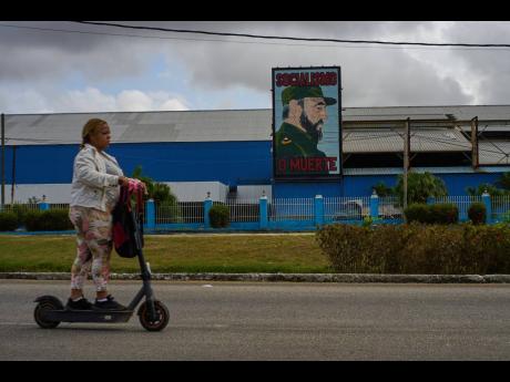 A woman rides an electric scooter past a factory displaying an image depicting the late Cuban leader Fidel Castro, bearing the words "Socialism or Death", in Havana, Cuba on March 19, 2026. (AP Photo/Ramon Espinosa)