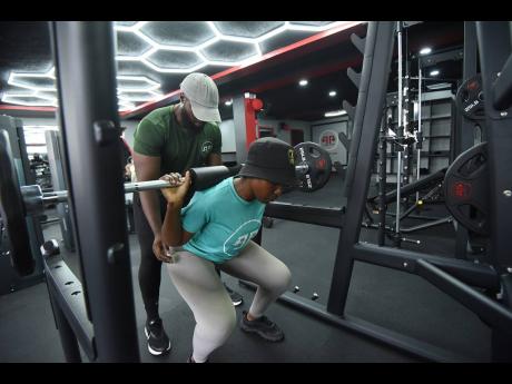 Samantha Francis performs a barbell squat while her personal trainer and husband, Rohan Francis, stands behind to provide support.